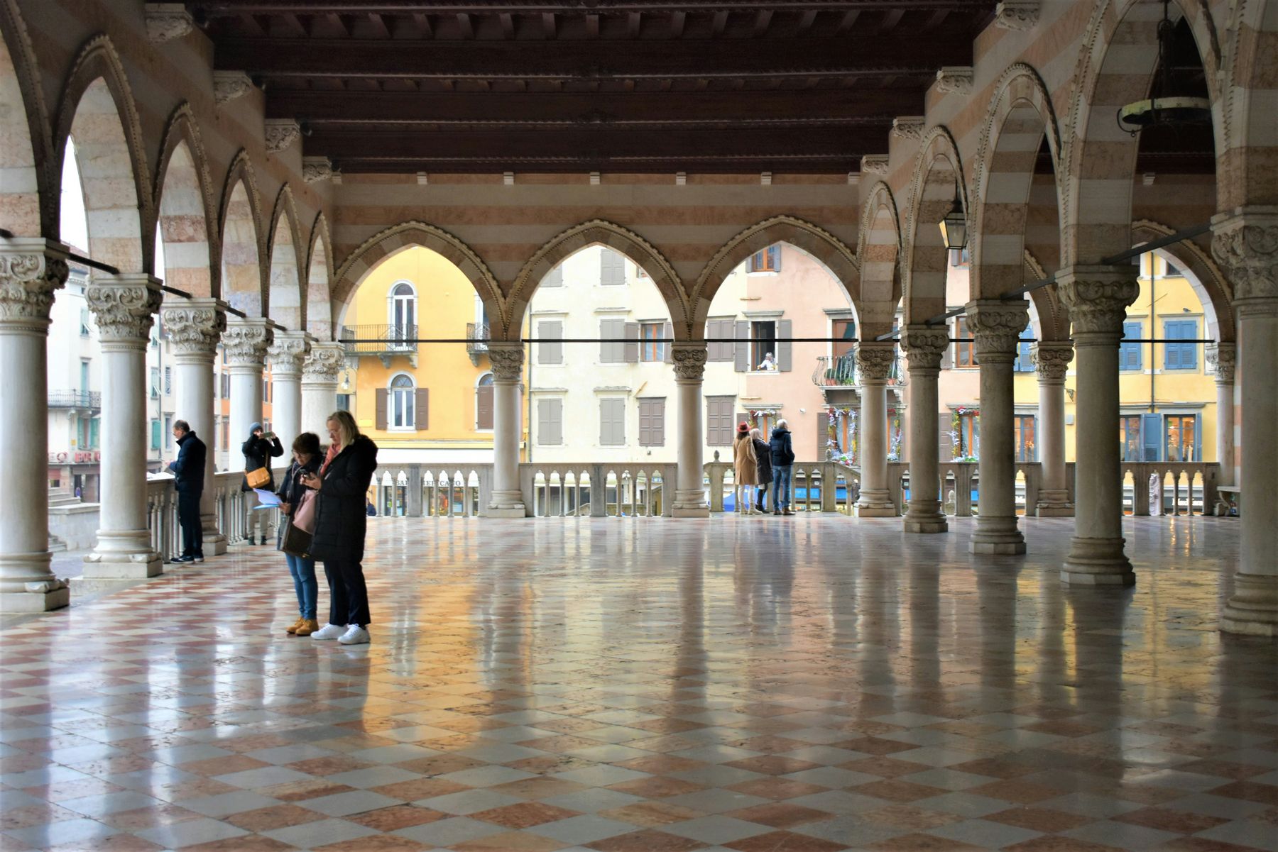 Sotto la loggia gotica del centro di Udine, con il pavimento a scacchi e la piazza oltre gli archi